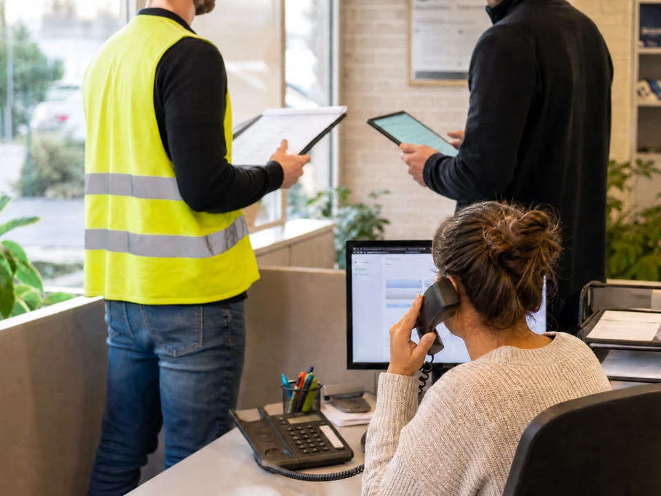 Photographie en intérieur d'une agence d'auto-école moderne. Un moniteur de conduite en polo écru discute avec une secrétaire au comptoir. Un bulletin psychotechnique ou dossier d'inscription visible sur le bureau. Ambiance accueillante et professionnelle, illustrant la diversité des rôles au sein de l'équipe.